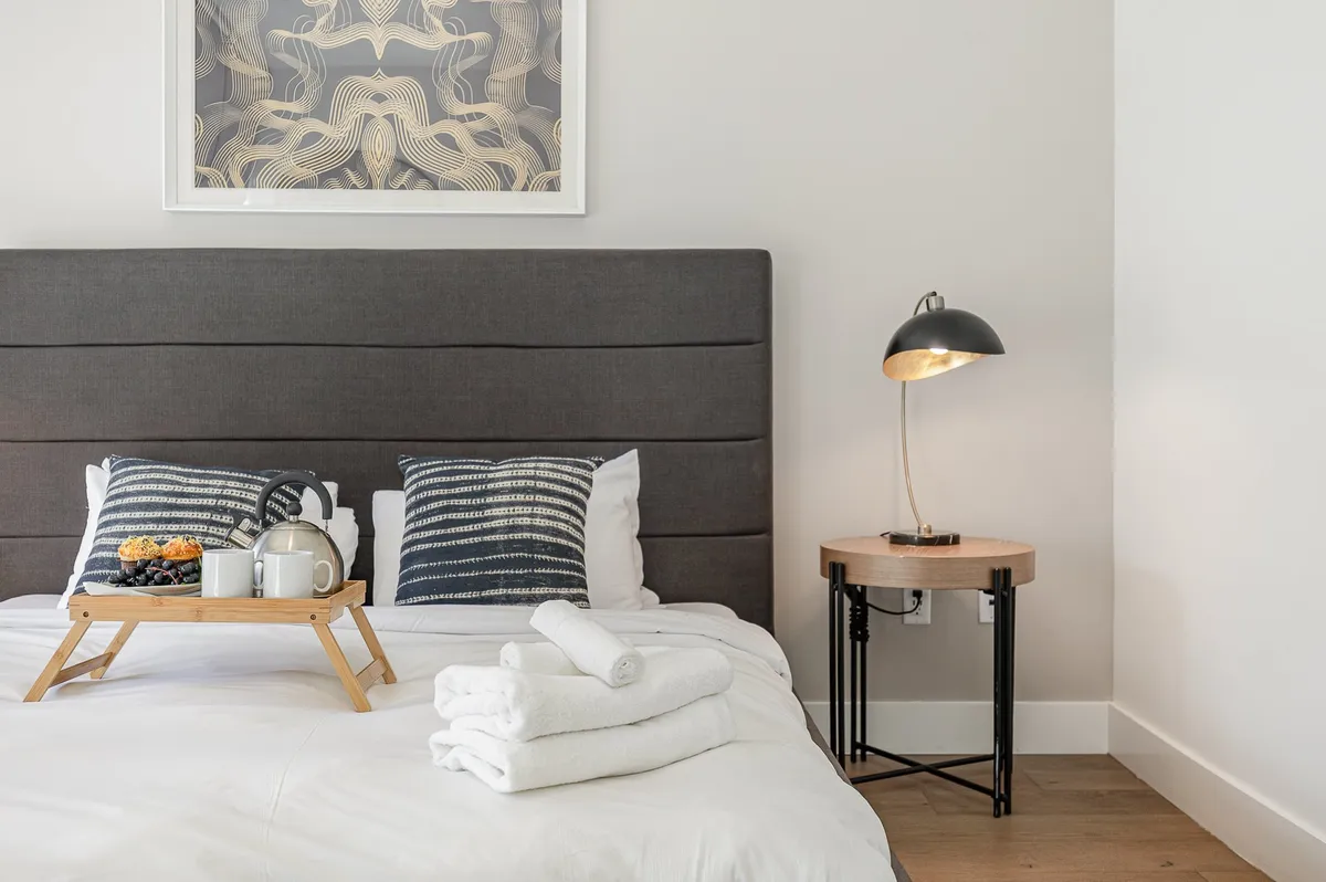 A cozy bedroom scene with a bed adorned with decorative pillows, a breakfast tray, soft lighting from a side lamp, and artwork above the headboard. The ambiance is serene and welcoming, ideal for relaxed mornings and quiet moments.