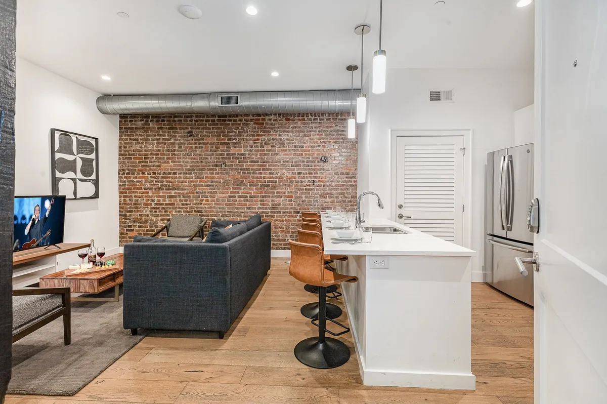 This interior scene highlights an inviting living room with a plush sofa, exposed brick wall, kitchen island, and television. The design is both modern and comfortable, making it perfect for relaxing or entertaining guests in style.