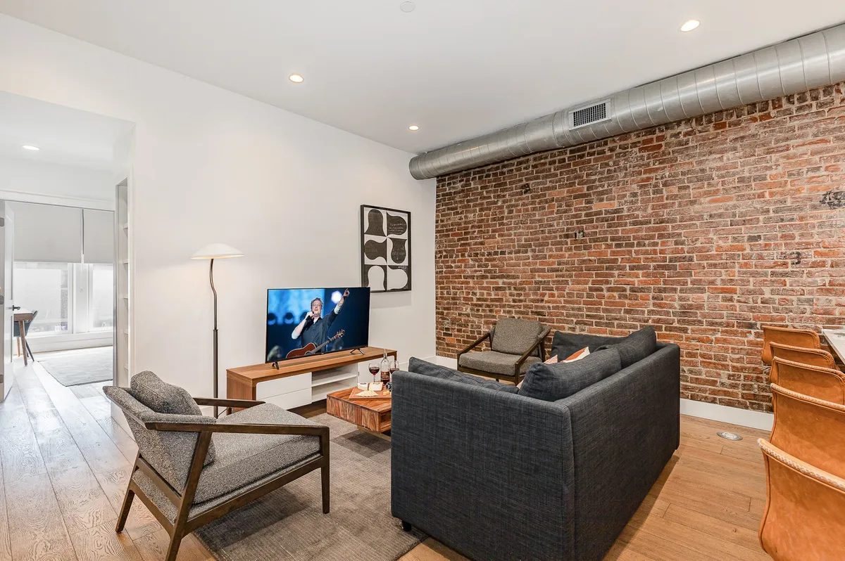 This image showcases a modern living room with a stylish exposed brick wall, a large television, plush sofa, armchairs, and a central coffee table. The space is arranged for both relaxation and socializing, with plenty of comfortable seating and a contemporary, inviting feel.