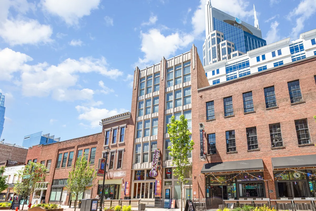 The image features the vibrant exterior of two historic brick buildings seamlessly blending into Nashville’s energetic cityscape, with modern skyscrapers towering above, lush green trees, and an inviting outdoor seating area perfect for socializing and relaxation. The setting is lively, suggesting proximity to music venues and bustling city life.