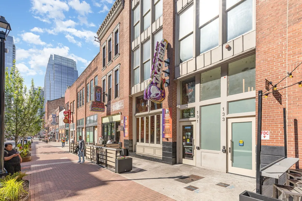 A lively street scene featuring brick facade buildings with visible restaurant signage, an outdoor seating area, and people socializing and exploring. The pedestrian walkway buzzes with activity, reflecting the vibrant local food and social scene that is key to a night out near Broadway.