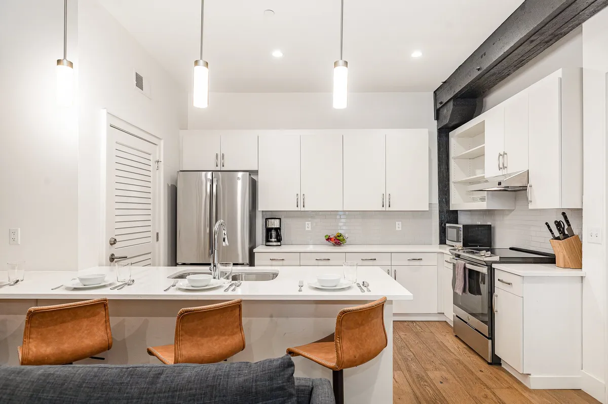 A bright, modern kitchen featuring a spacious island with bar stools, gleaming stainless steel appliances, and crisp white cabinetry. The space is designed for social cooking, entertaining, and sharing meals, with plenty of room for group activities and celebrations.