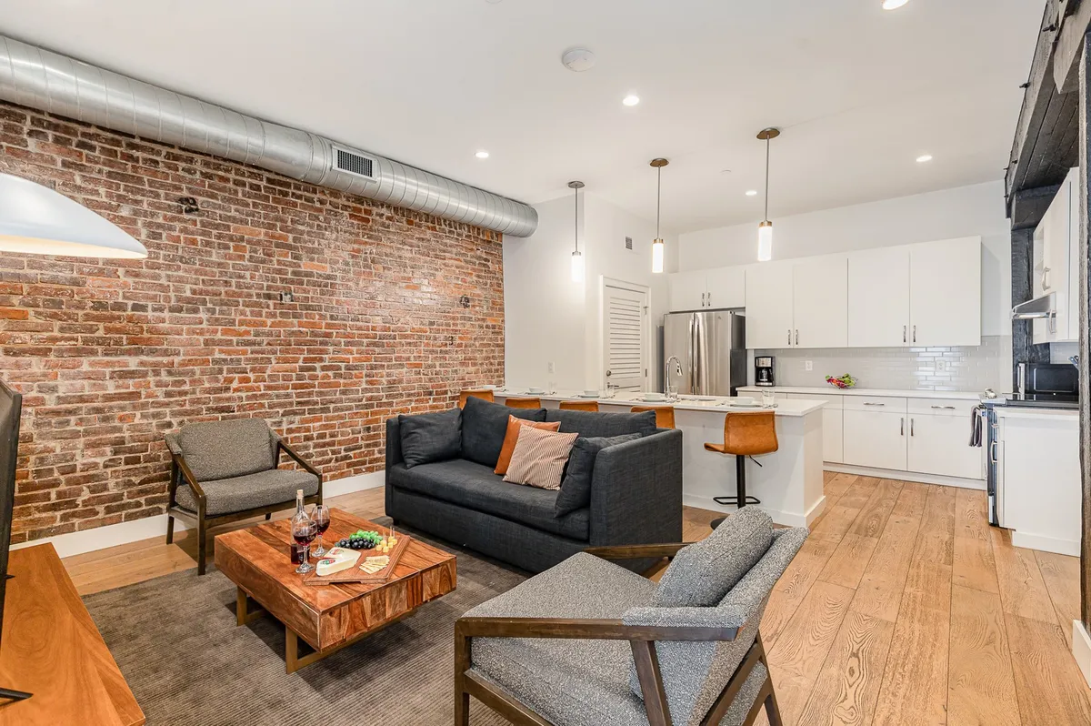 A cozy and inviting living space featuring a modern sofa, exposed brick wall, wooden coffee table, and a kitchen island visible in the background. Warm lighting and a layout that encourages conversation and group gatherings are evident.