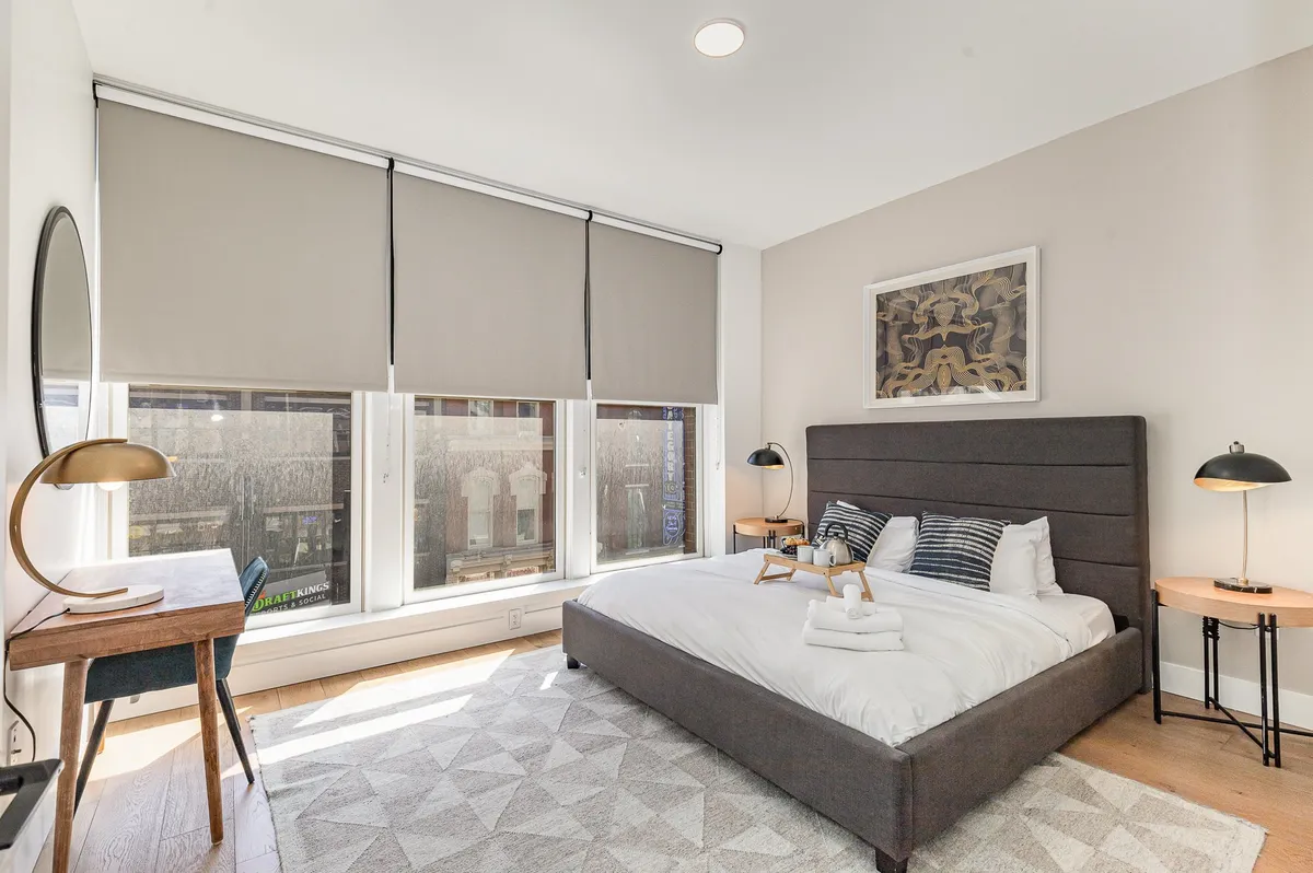 A peaceful bedroom with a comfortable bed, large windows letting in natural light, a functional desk for work or study, and artistic touches on the wall. The space blends rest, productivity, and visual appeal.