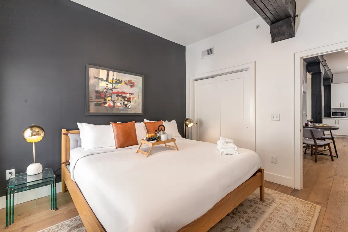A stylish bedroom featuring a plush bed with decorative pillows, large wall artwork, a golden lamp, and a tray with refreshments set on the bed. The lighting and textures create a cozy, inviting atmosphere ideal for relaxation.